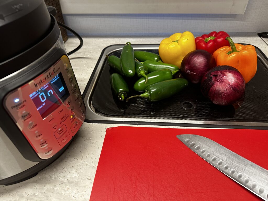 instant pot on left and uncut peppers, onions, and jalapeños on counter with cutting board and knife.