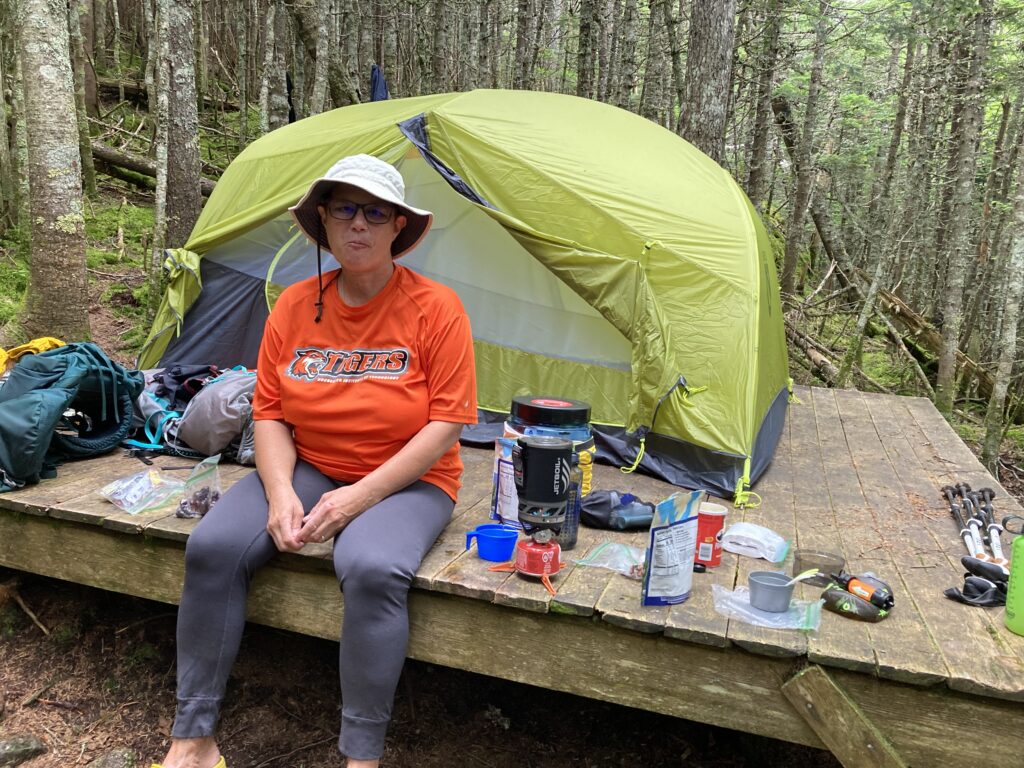 woman hiker sitting in front of tent on platform in woods with backpacking gear spread out around her