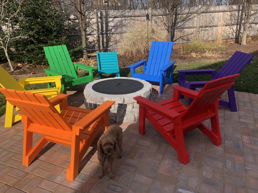 dog next to multi colored Adirondak chairs on patio around fire pit.